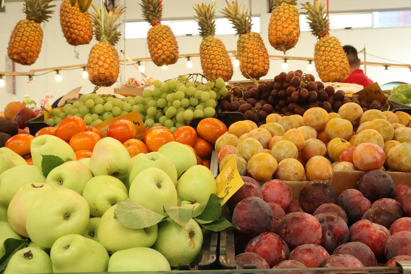 Fruta em exposição na banca da Cesta Cheia, com maçãs, uvas, ameixas e ananases pendurados